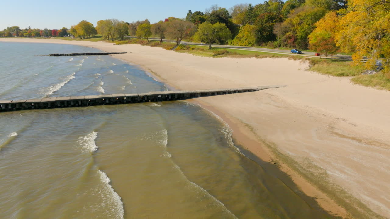 Drone aerial over the Lake Michigan shoreline in Sheboygan, Wisconsin drifting down toward a pier with gentle waves and colorful autumn scenery on a clear day