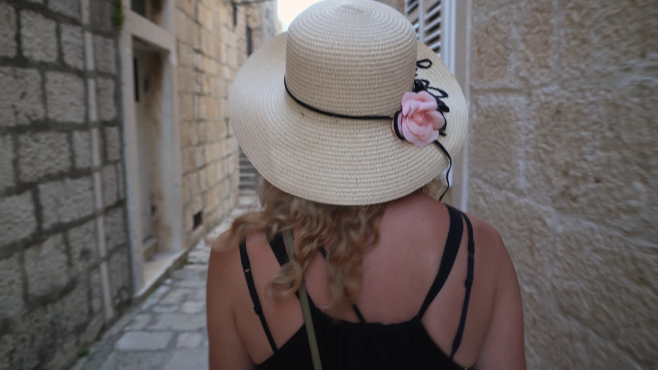 Caucasian Female Tourist Wearing A Summer Hat Is Walking On The Alley Of Historic Town In Croatia. Tracking Shot