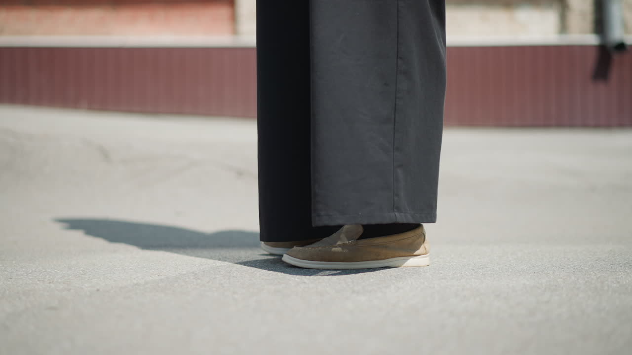 Person stands quietly on sunlit pavement, wearing black wide pants and brown shoes, strong light casts clear shadow, suggesting waiting, contemplation in calm outdoor environment