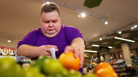 An-overweight-man-wearing-a-purple-T-shirt-chooses-oranges-from-the-counter-and-puts-them-in-a-bag-in-a-supermarket.-Daily-routine-of-an-overweight-man-in-a-supermarket.-Shopping-trip