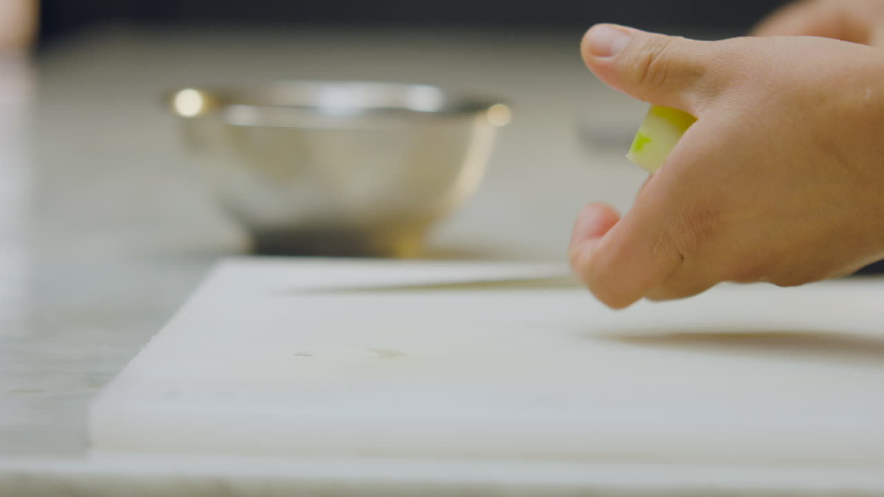 Close-up of a chef's hands peeling a green apple with a peeler on a white chopping board, next to a knife and an aluminum mold on a white countertop