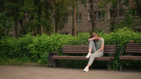 young woman sitting on wooden bench in outdoor park setting, resting her head on her arm in a reflective and somber posture, surrounded by lush greenery and trees