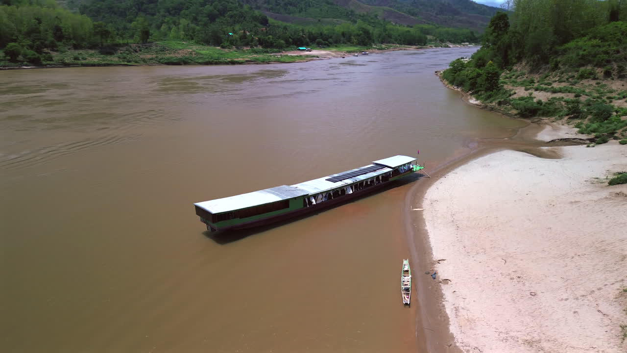 Aerial orbit of slow boat gliding down brown Mekong River in rural Southeast Asia approaching sandy banks