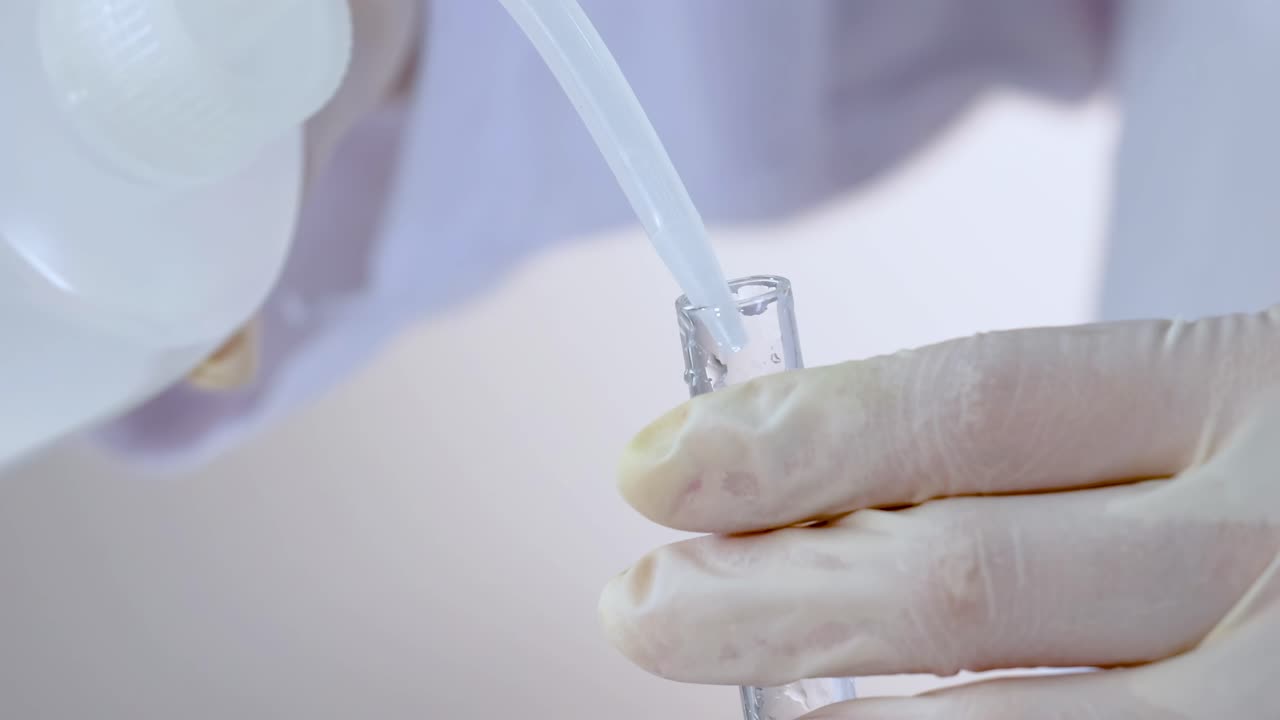 Close-up of gloved hands pouring liquid from a bottle into a test tube.