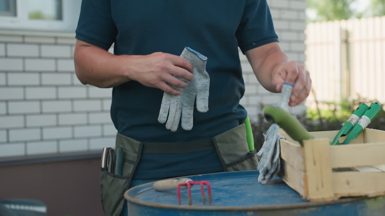 Close up gardener drops trowel fork on blue drum, picks gloves from wooden crate beside house, apron belt around waist visible, summer yard scene