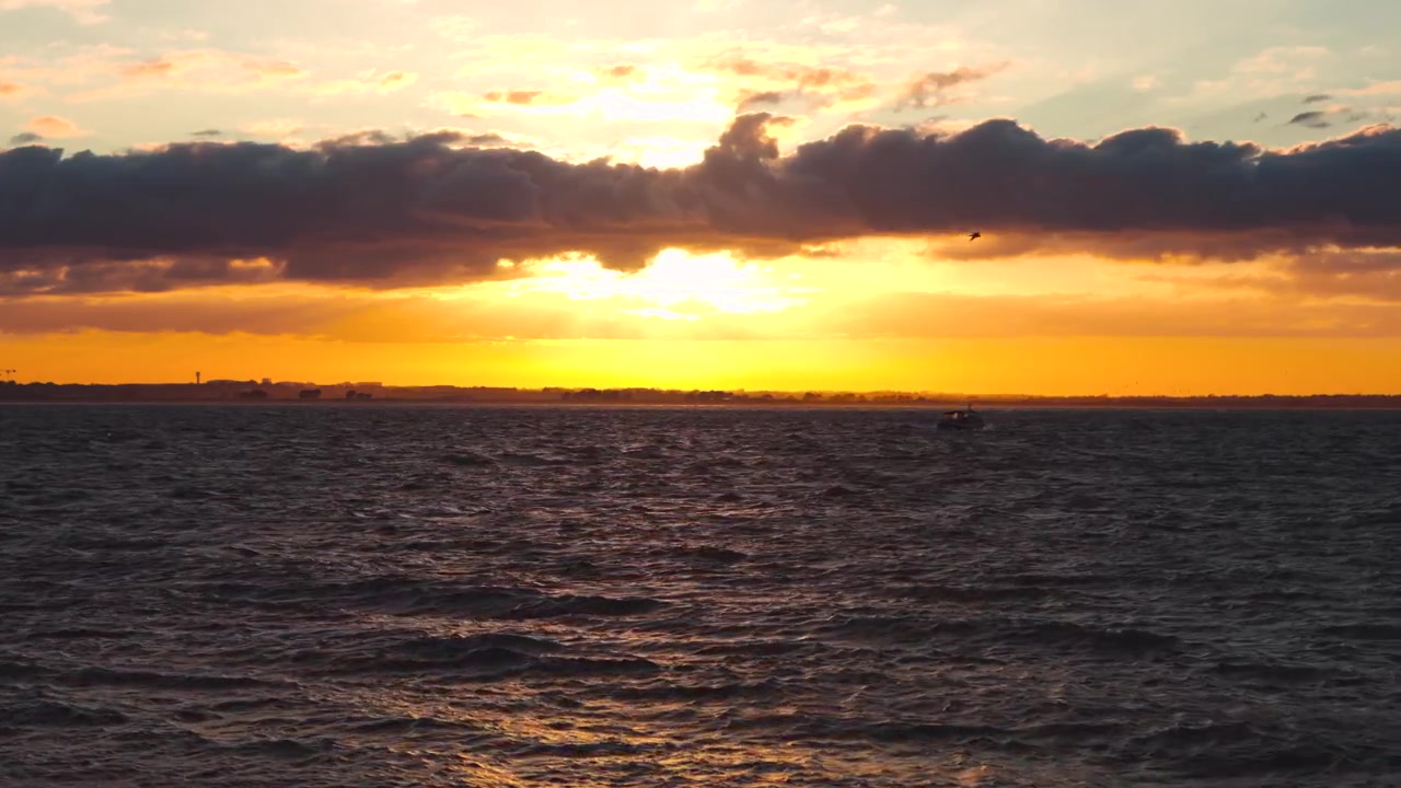Distant boat crossing rough waters beneath a golden sunset as clouds cover the evening sky