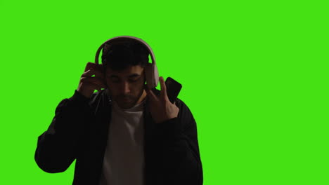 Man-Putting-On-Wireless-Headphones-And-Streaming-Music-From-Mobile-Phone-Against-Green-Screen-With-Low-Key-Lighting-1