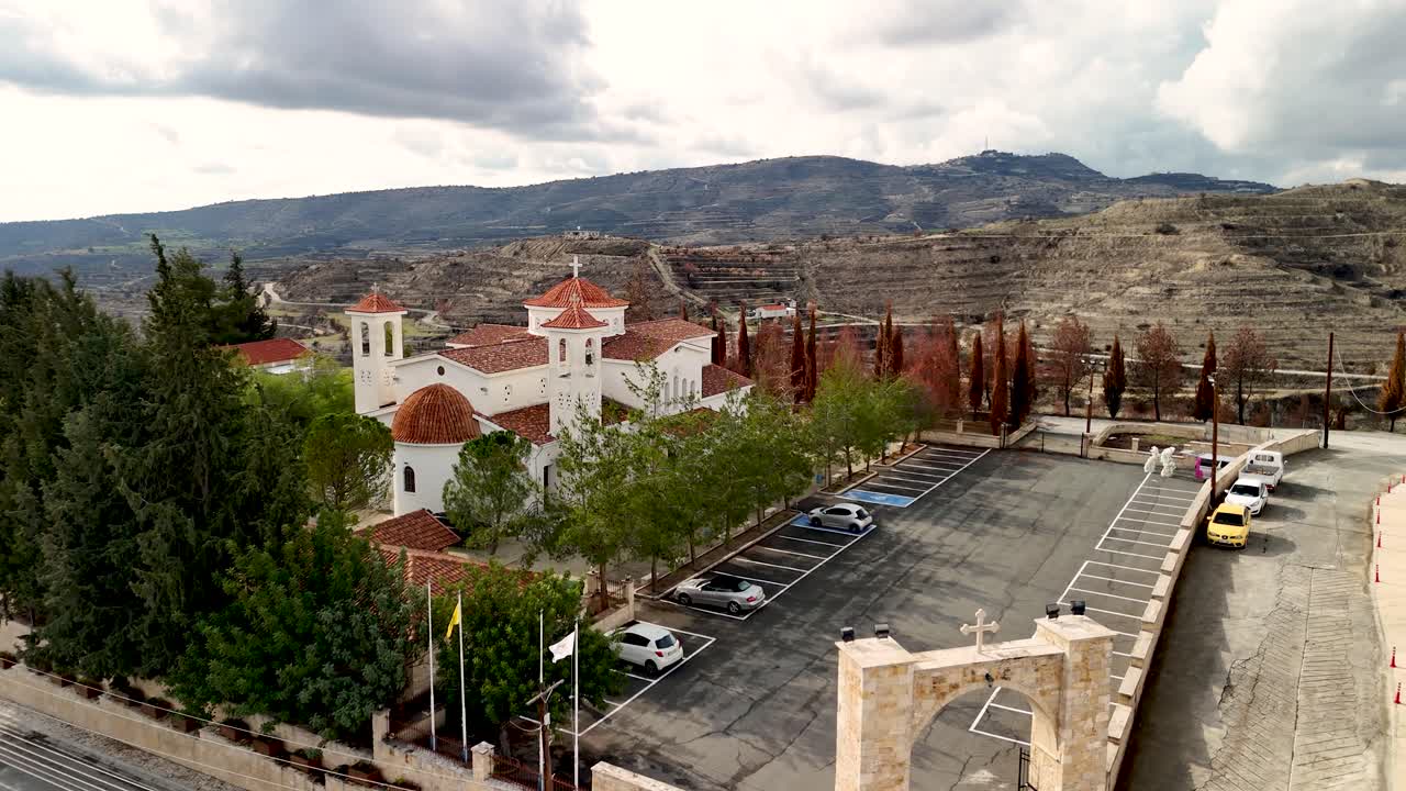 Una chiesa si erge tra alberi alti e vigneti sulle colline di Cipro. Nuvole coprono il cielo. Le auto parcheggiano vicino all'edificio. Il paesaggio mostra un mix di natura e strutture.