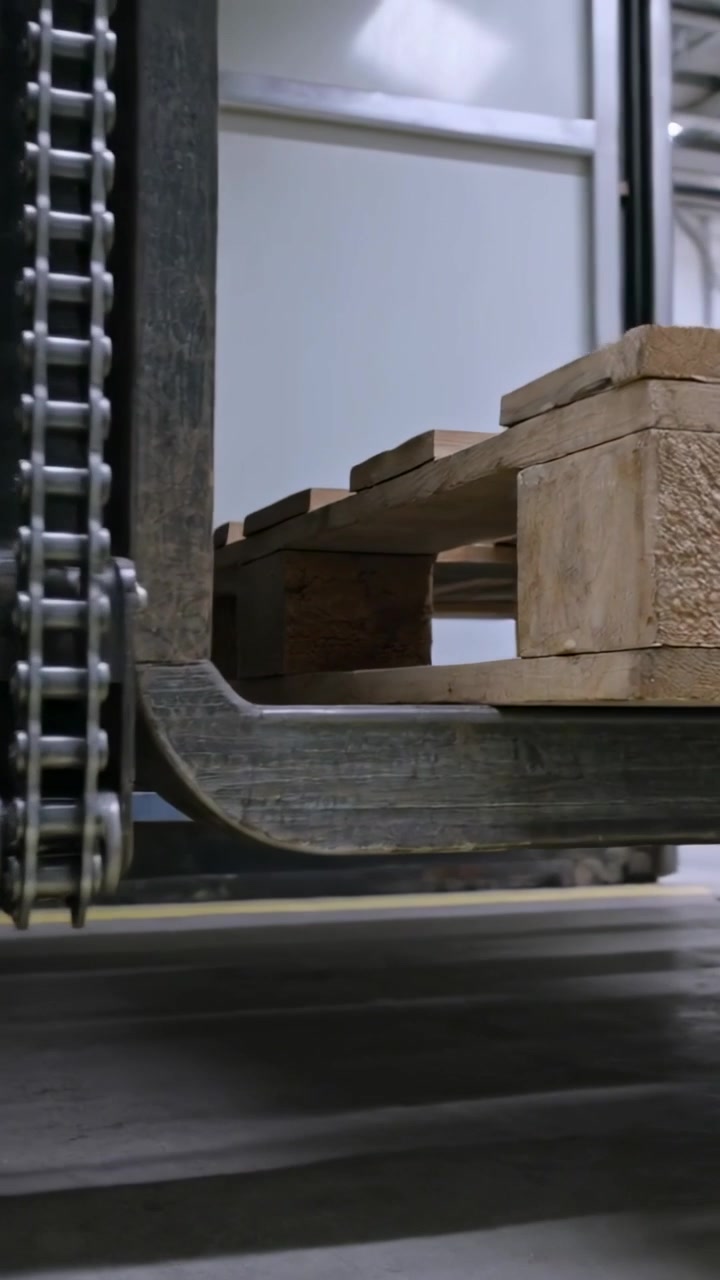 Video verticale: Denti del carrello elevatore che scorrono sotto un pallet di legno in un magazzino, con il sollevatore che si attiva per caricare.