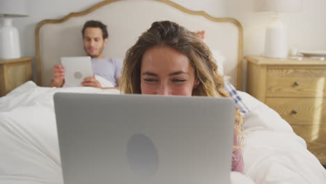 couple wearing pyjamas lying in bed at home with woman using laptop and man digital tablet