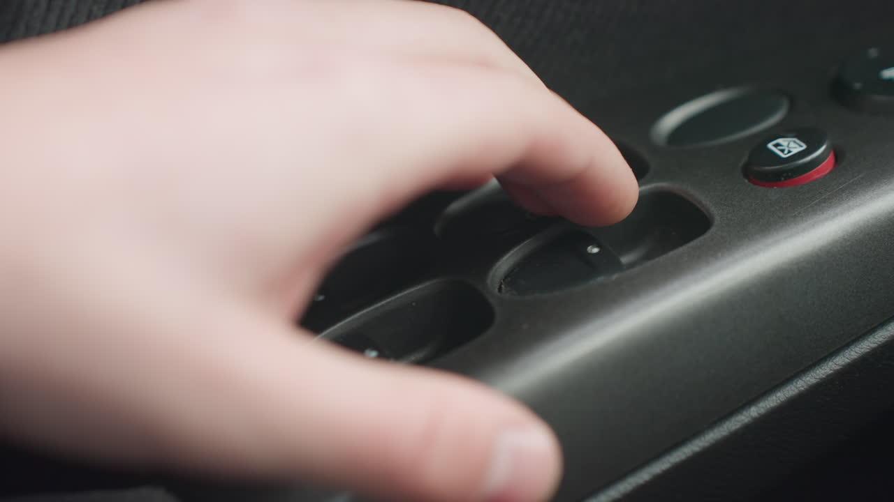 close up detailed view of window control buttons on car door panel as fair skinned hand reaches toward auto switch showing texture of plastic surface and dark fabric interior