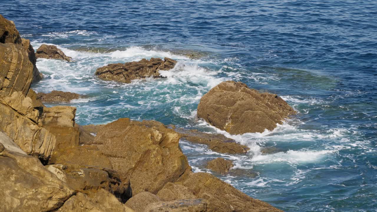 Rough, multi-layered brown rocks on the San Sebastian coast with turquoise waves breaking