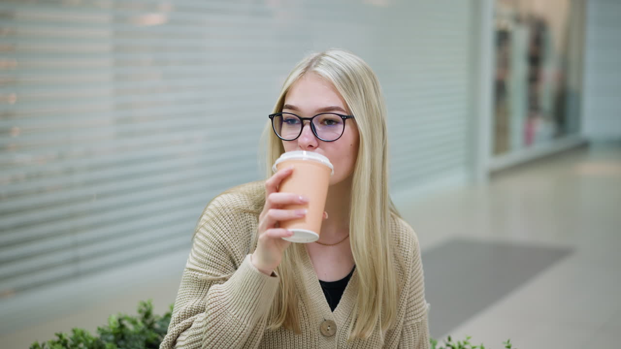 una joven sentada en un centro comercial bebiendo una bebida mientras mira hacia otro lado, con una tienda en el fondo mostrando bolsas y mercancías coloridas