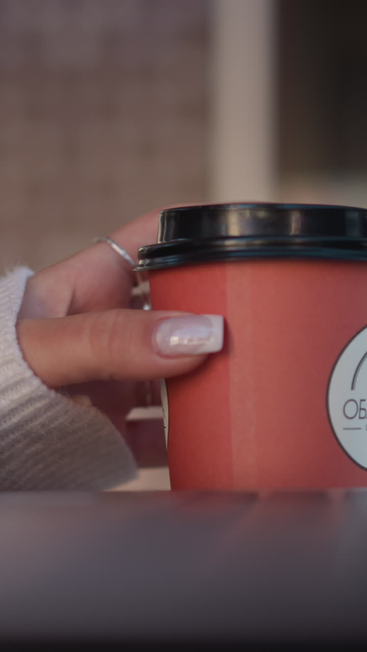 primer plano de la mano de la dama girando suavemente la taza de café cerca de la computadora portátil con enfoque en los dedos manicuñados, la mano de escritura borrosa y la silla en el fondo