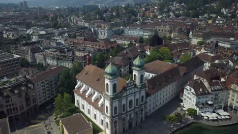 órbita-Aérea-De-La-Iglesia-Jesuita-Barroca-En-El-Casco-Antiguo-De-Lucerna-Junto-Al-Río-Reuss,-El-Puente-De-La-Capilla-Y-La-Torre-De-Agua,-Colinas-En-El-Fondo,-Suiza