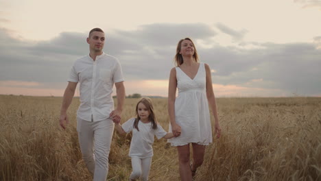 Young-couple-of-parents-with-girl-children-holding-hands-of-each-other-and-running-through-wheat-field-at-sunset.-Happy-family-jogging-among-barley-meadow-and-enjoying-nature-together.-Slow-motion