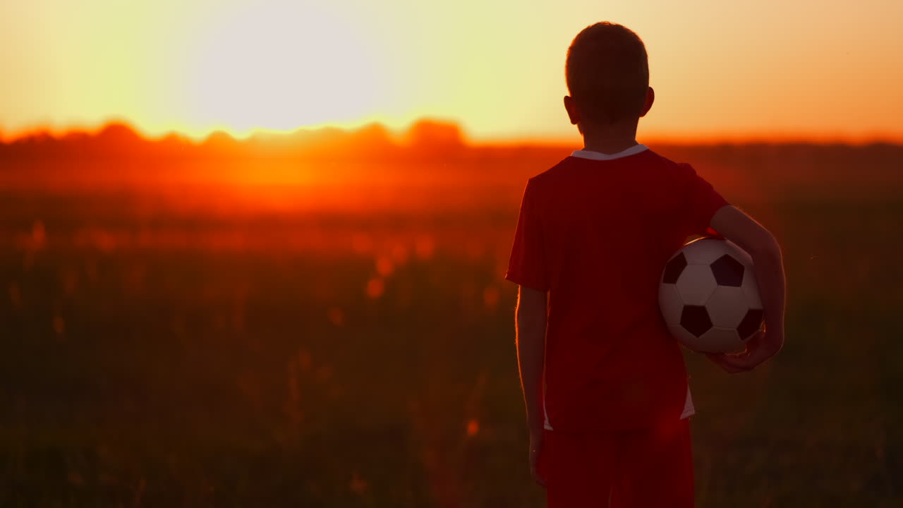 un niño con una pelota en un campo al atardecer. un niño sueña con convertirse en un jugador de fútbol. un niño va al campo con la pelota al anochecer.
