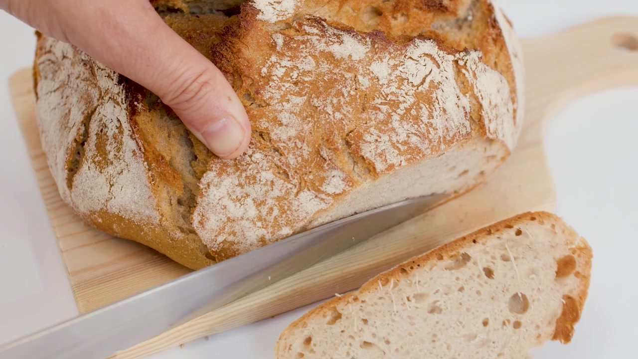Primo piano di mani che tagliano una pagnotta di pane a lievitazione naturale croccante su un tagliere di legno su sfondo bianco.