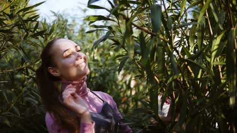 feliz mujer jardinera sonriente en delantal examinando las hojas de diferentes árboles mientras camina entre filas de árboles en un jardín o invernadero