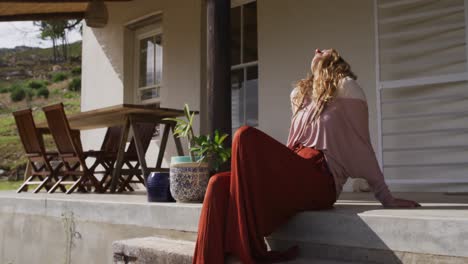 Feliz-Mujer-Caucásica-Sentada-En-La-Soleada-Terraza-De-La-Cabaña,-Relajándose-Con-Los-Ojos-Cerrados