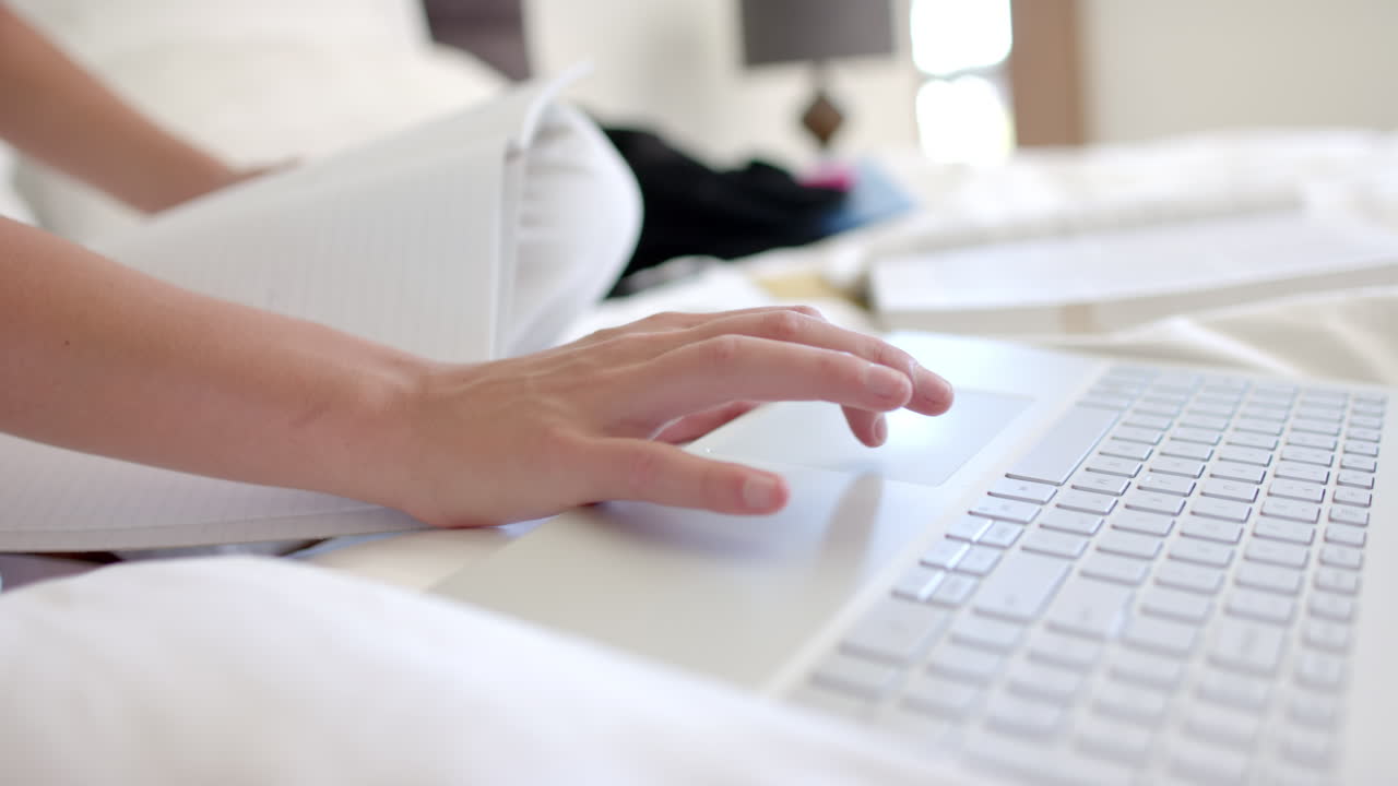 Close-up of a teenage girl''s hands typing on a laptop with a notebook beside them at home