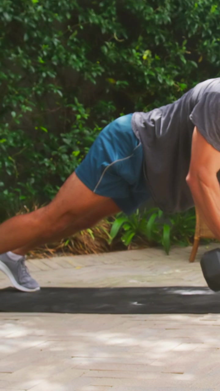 Video verticale: Uomo che si allena in pantaloncini, facendo flessioni e rematore con manubri su un patio per la forza.