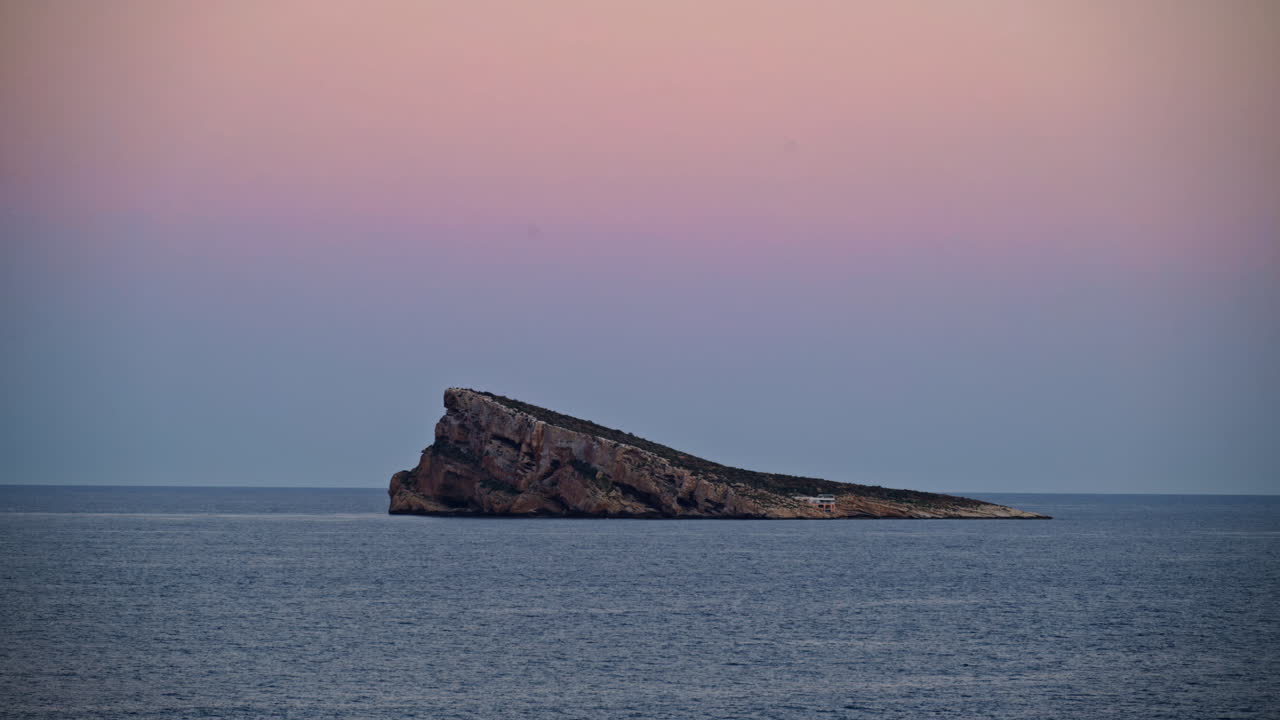 Dusk falling over a lonely offshore islet as a gull glides through a pastel sky in Benidorm, Spain
