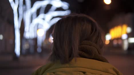 Beautiful-tender-girl-is-walking-through-night-city-in-coat.-Turns-around-and-smiling.-Tracking-shot