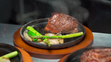 Steak-beef-prime-picanha-tbone-new-york-on-iron-plates-steam-hot-with-onion-and-asparagus-tilt-shot-on-restaurant-kitchen-wooden-base-plate