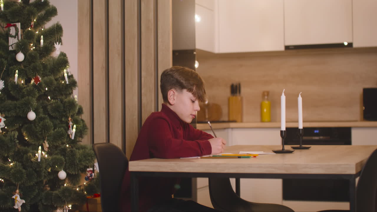 Side View Of A Boy In Red Turtleneck Writing A Letter Of Wishes Sitting At A Table In A Room Decorated With A Christmas Tree 1