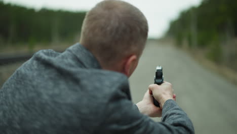 a close-up view from behind of a man in a gray jacket aiming a handgun down a deserted railway track, having blurred forest background