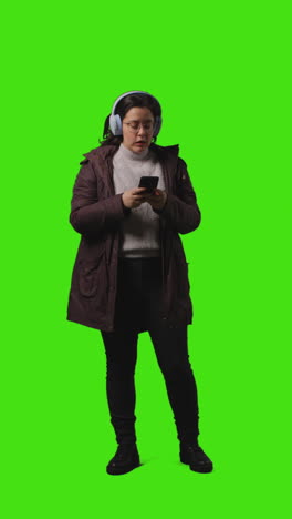 vertical video full length shot of woman wearing wireless headphones streaming music from mobile phone walking into frame against green screen