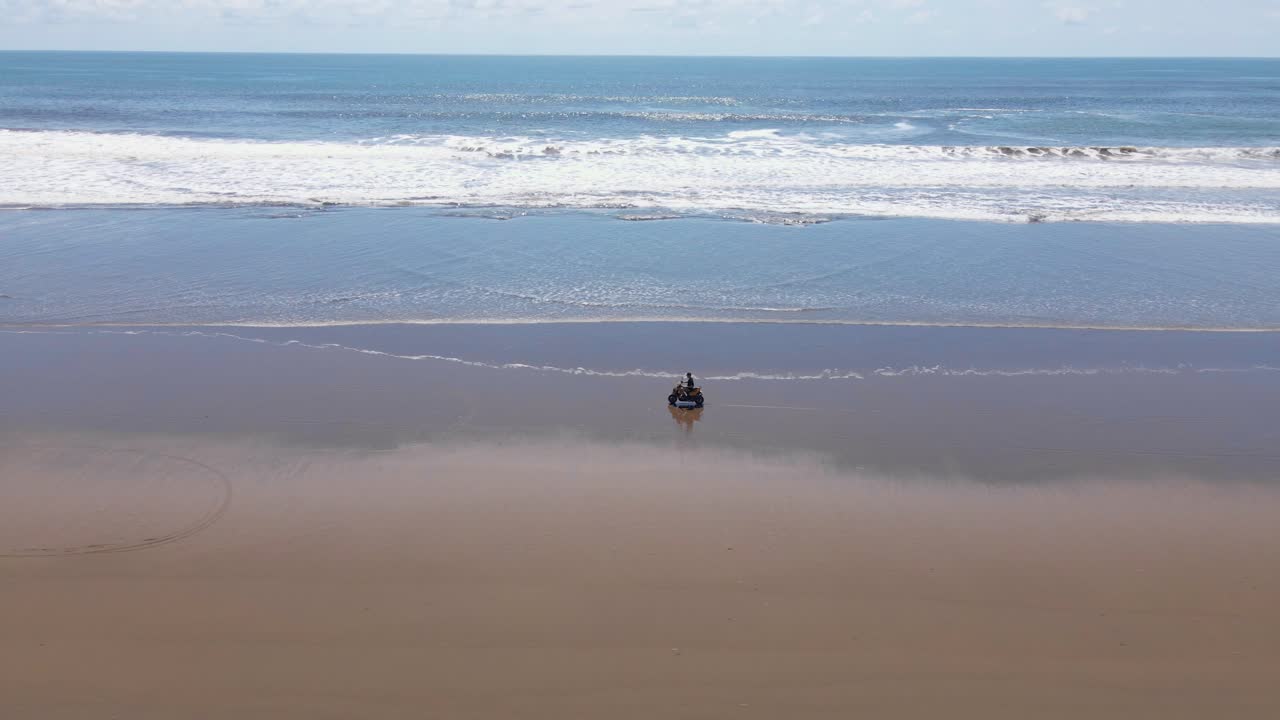 햇빛 아래 빈 해변을 따라 달리는 동안 스쿠터를 타고 사람을 옆으로 추적하는 드론