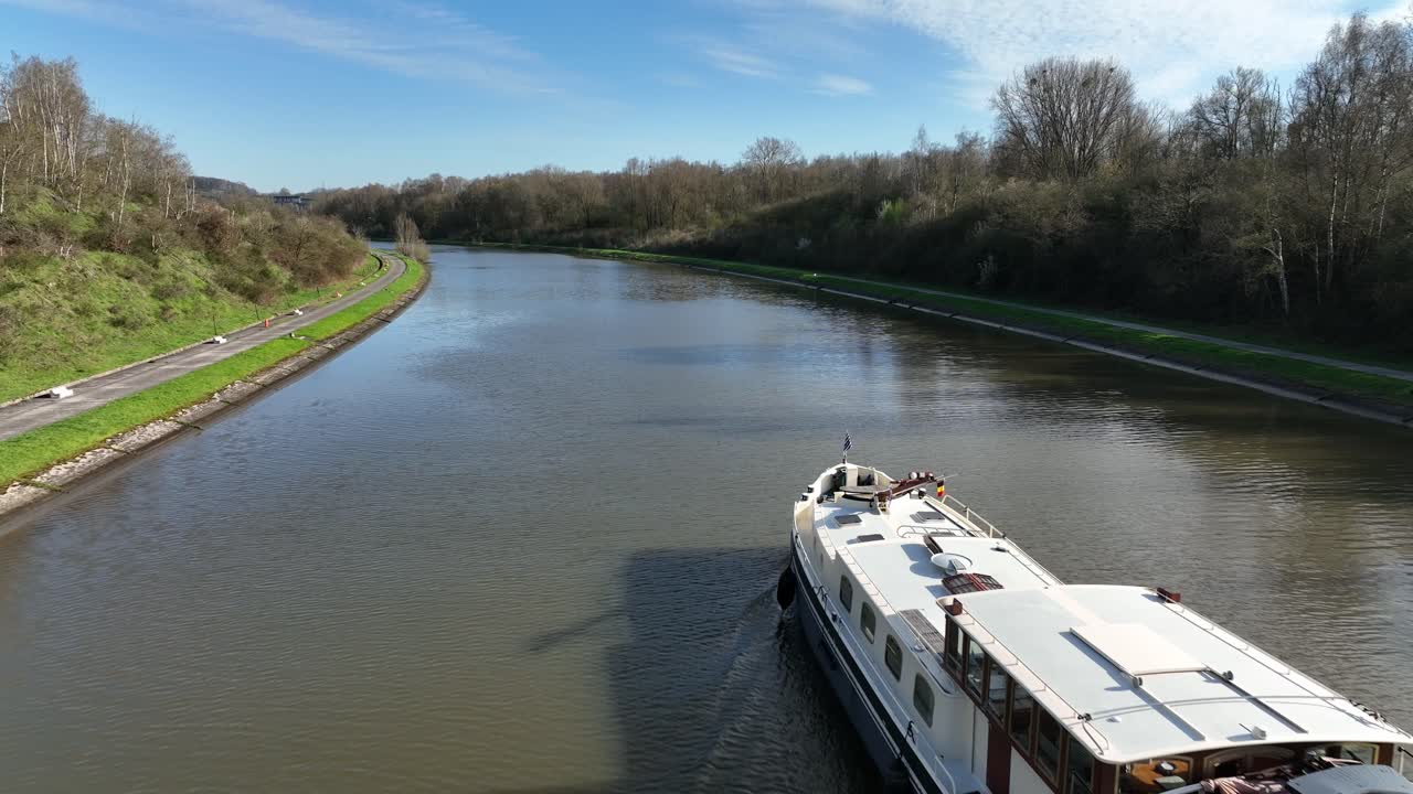 Aerial zoom in towards a boat navigating canal in Brussels, Belgium, surrounded by trees and fields under sunny skies
