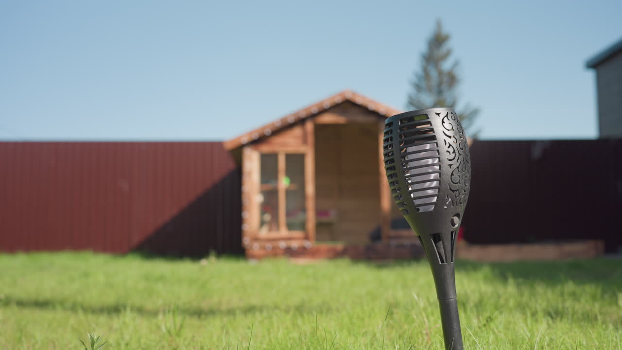 Wooden kids playhouse with glass panels placed on fresh green grass in sunny backyard, accompanied by small sandbox, tall pine tree, red metal fence, and blurred garden light in foreground