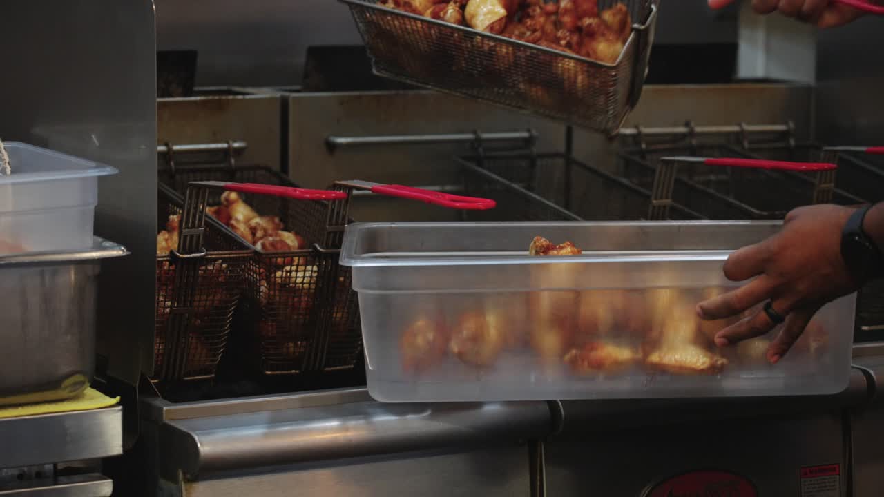 Filmato 4K di ali di pollo appena fritte che vengono versate in un grande contenitore dopo la cottura in una friggitrice professionale, catturando la consistenza calda e croccante e la preparazione del cibo in una cucina commerciale