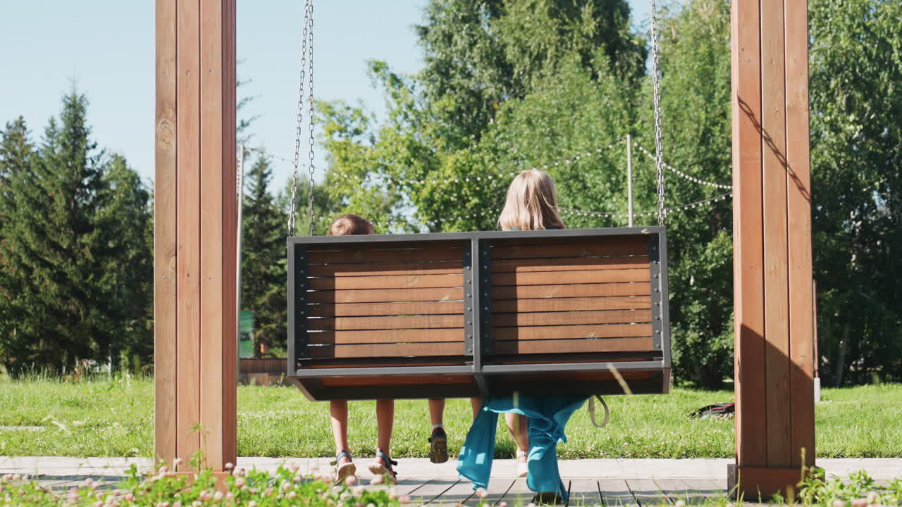 Barn og forælder på gynge under lyse sommertræer, blå kjole og svingende ben, glad pegende gestus og latter, sollys på græs og ubekymret weekendenergi, der fanger den fantasifulde udendørs stemning.