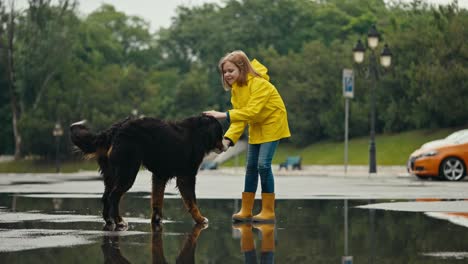 Happy-teenage-girl-in-a-yellow-jacket-stands-in-a-puddle-in-rubber-boots-and-pets-a-large-black-dog-while-walking-in-the-park-after-the-rain