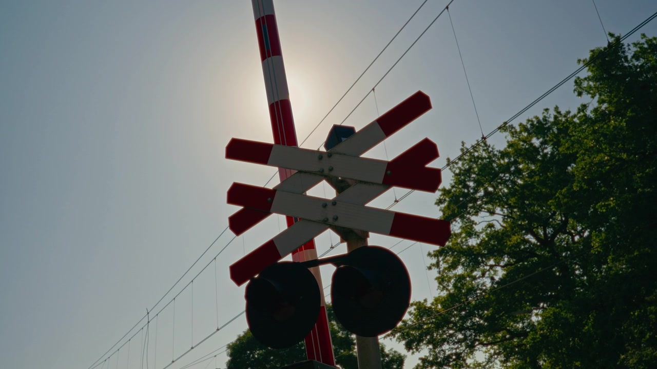 Close up of a red and white railway crossing signal, warning lights and raised barrier against a clear blue sky, with overhead railway power lines and a subtle sun flare.