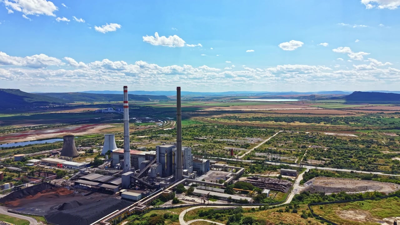 Usina de energia localizada em uma área industrial perto de um rio, cercada por campos e colinas sob um céu claro com nuvens