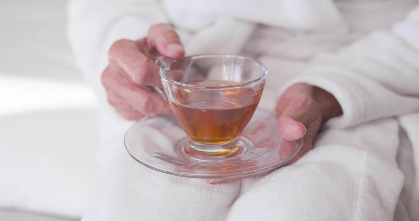 Donna afroamericana anziana che si sistema in vestaglia, tenendo una tazza da tè e un piattino in grembo a casa, per stabilizzare la tazza.