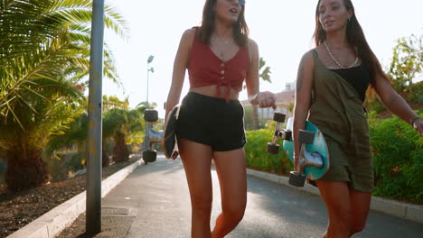 in the backdrop of the setting sun, girlfriends carrying skateboards walk near palm trees, engaged in conversation, laughter, and joyful smiles, the scene captured in slow motion