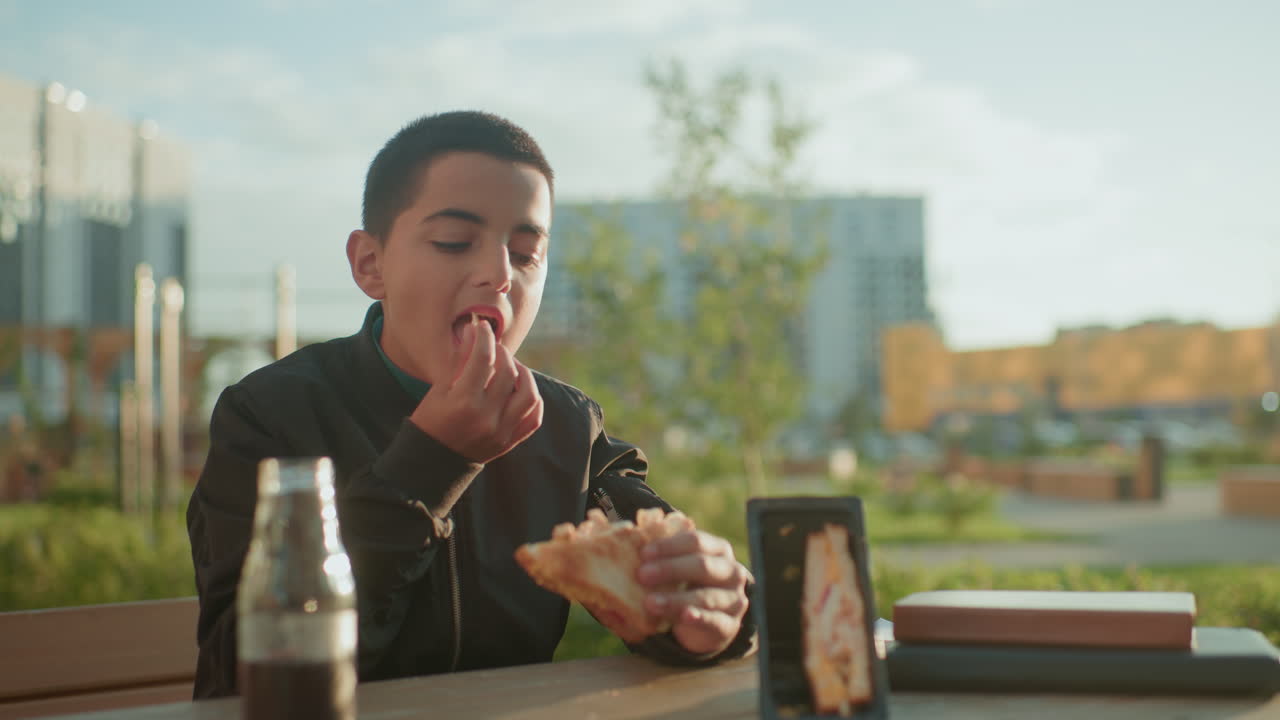 Boy sitting outdoors eating pizza with juice bottle and sandwich pack on wooden table, enjoying snack in warm sunlight with relaxed expression, casual atmosphere in modern urban background