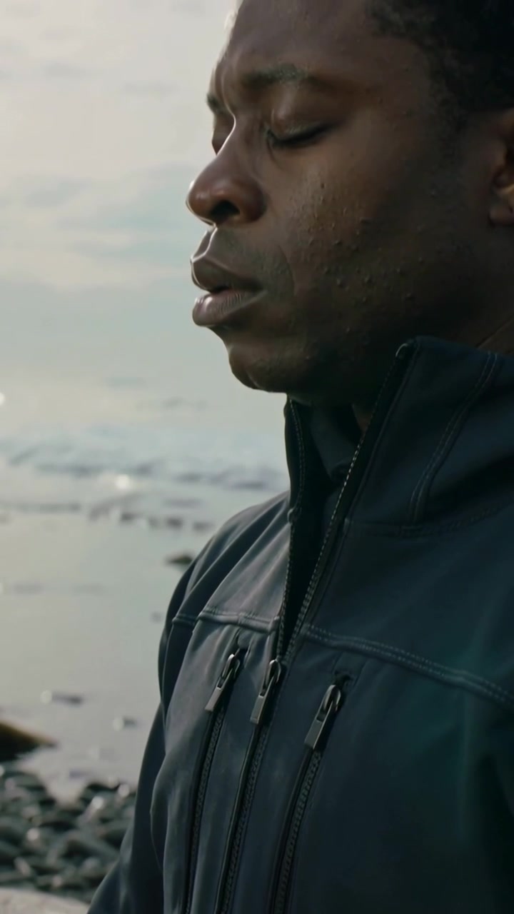 Vertical video: Camera lifting, man closing eyes, reflecting on rocky shore wearing dark zip jacket
