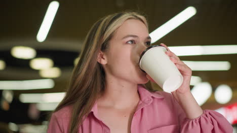 lady in pink dress seated in restaurant takes a sip of coffee from her cup with a satisfied expression, nodding in approval while enjoying the taste, the background shows blurred lights and people