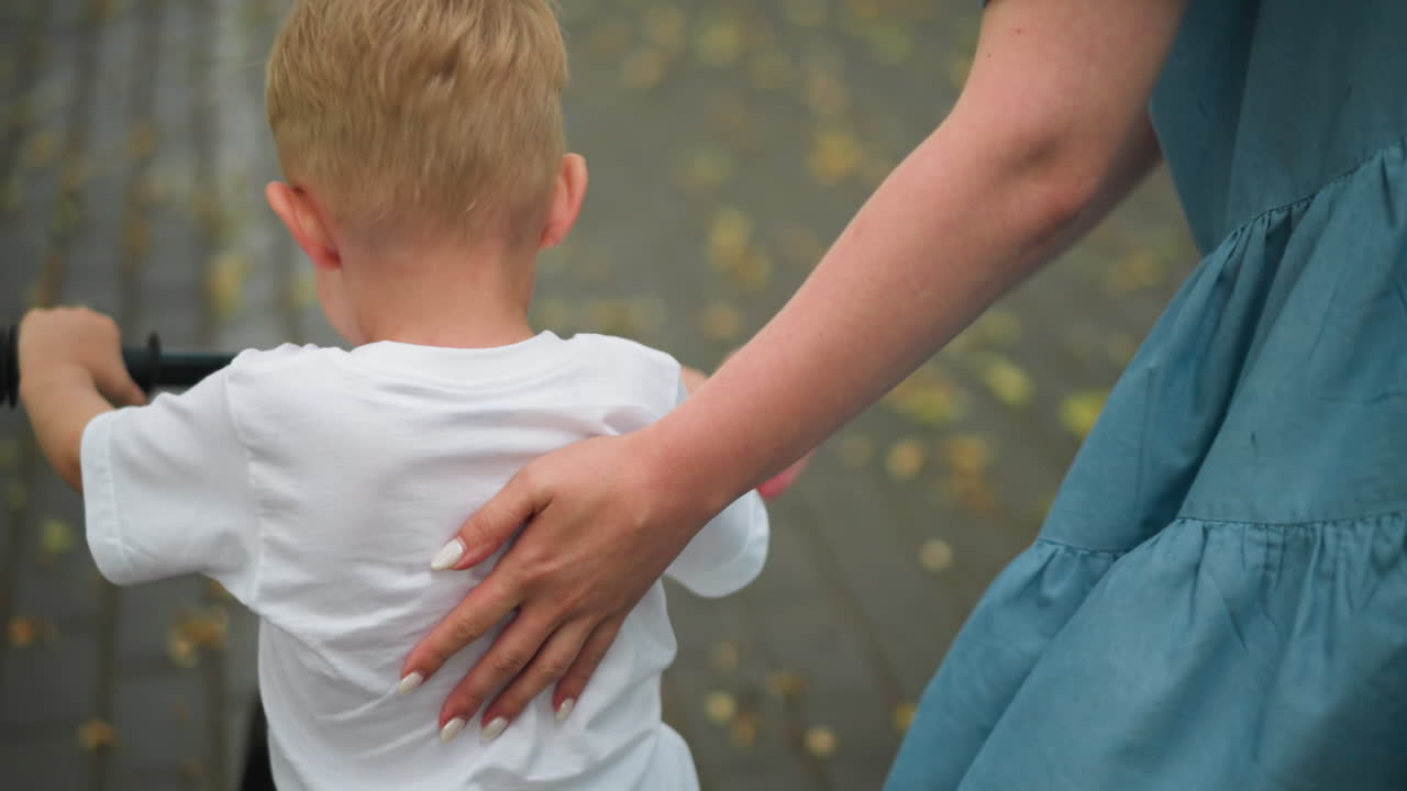 una madre con uñas pintadas apoya suavemente a su hijo pequeño aprendiendo a montar una bicicleta, el niño usa su pierna para equilibrarse mientras avanzan por un camino sembrado de hojas