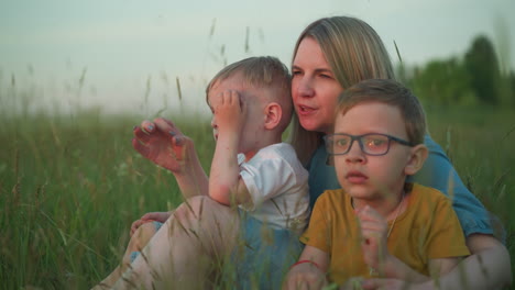 close-up shot of a mother in a blue gown sitting in a field with her two children, one wearing a yellow top and glasses, and the other in a white top.they gently touch the grass and gaze at the nature