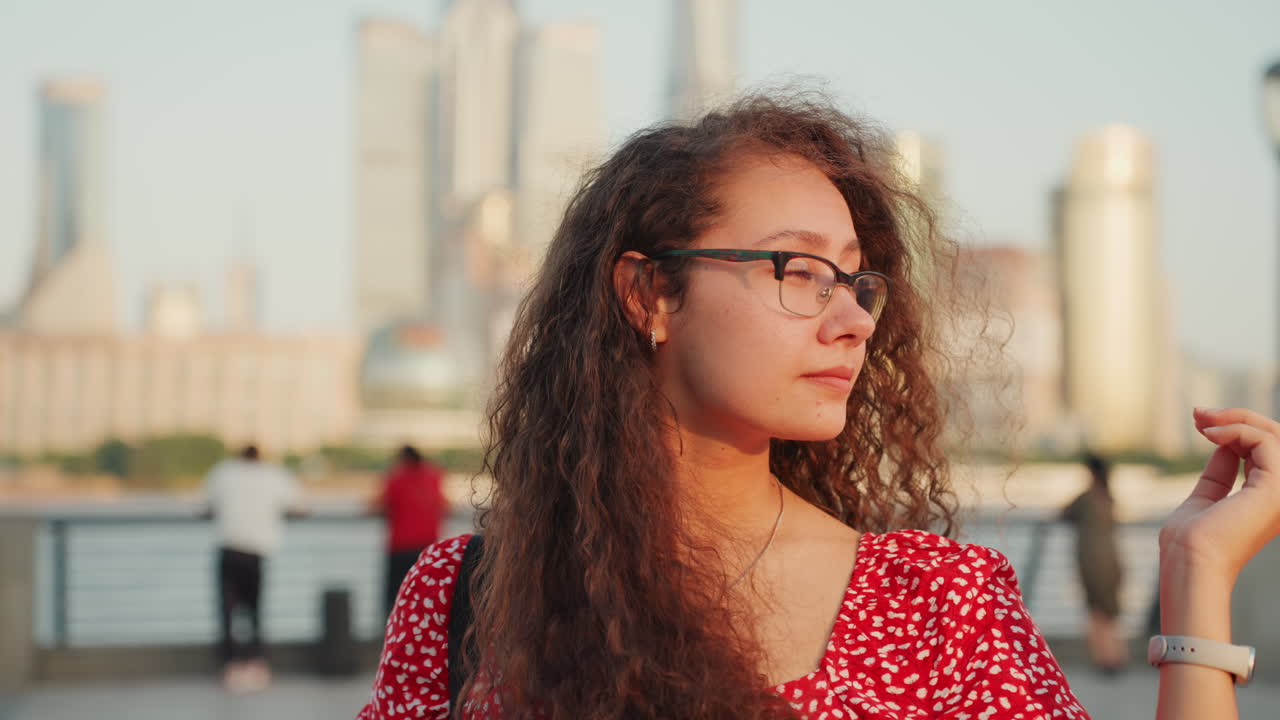 Giovane donna in abito rosso sta in piedi sul lungofiume all'ora d'oro, con gli occhiali e i capelli ricci mossi dalla brezza, lo skyline della città sfocato al di là del fiume, si aggiusta gli occhiali e scruta l'orizzonte.