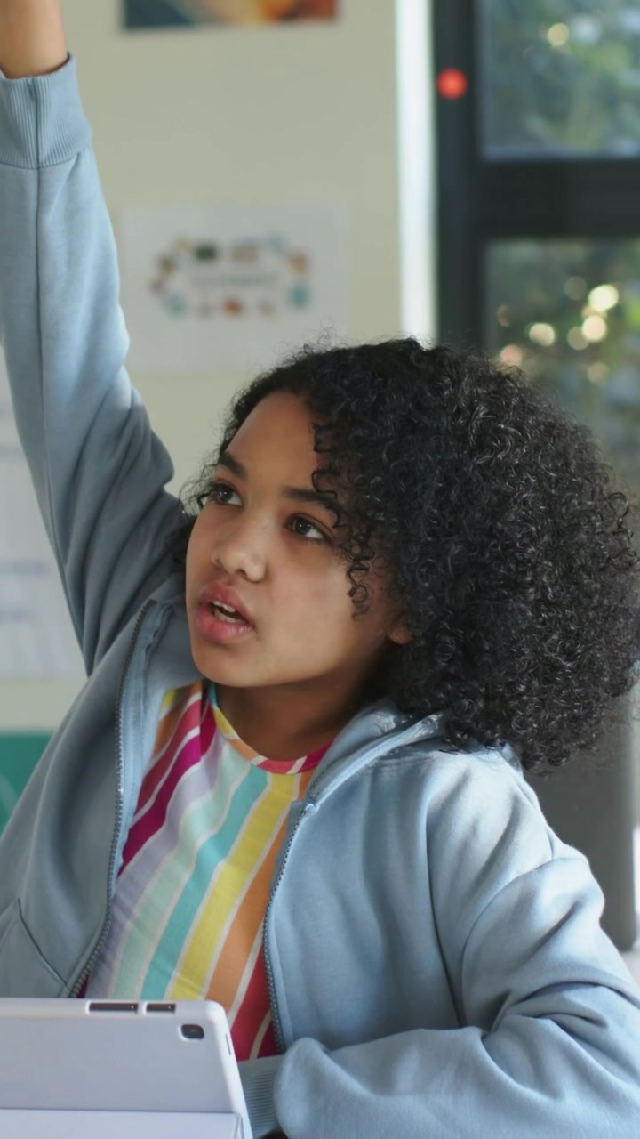 Video verticale: Notando l'indicazione dell'insegnante, una ragazza con felpa blu che alza la mano destra in classe, tablet