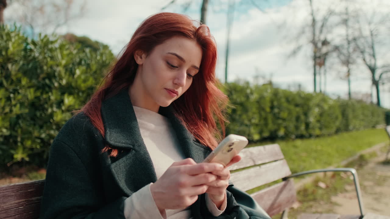 Giovane donna dai capelli rossi che usa lo smartphone su una panchina del parco